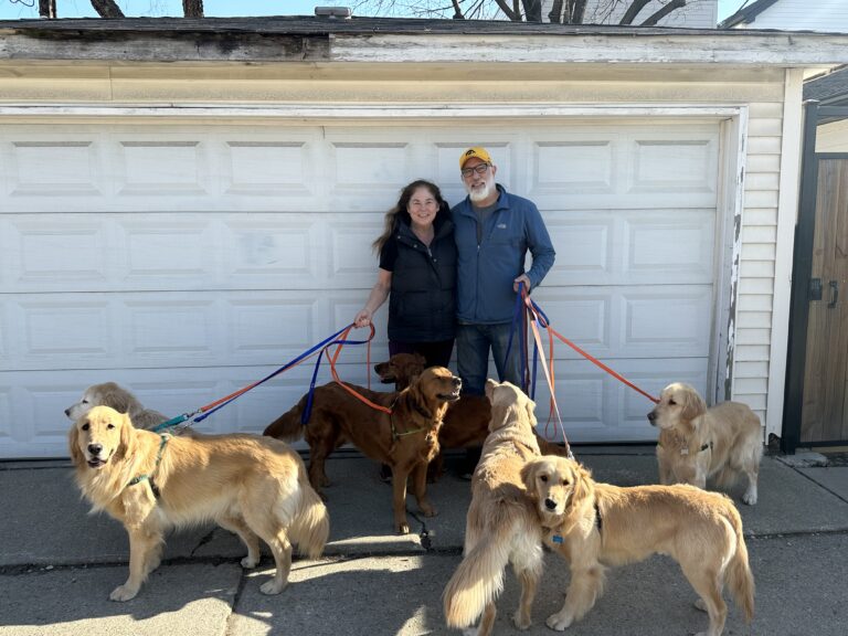 smiling couple in front of a garage door with 5 dogs on leashes